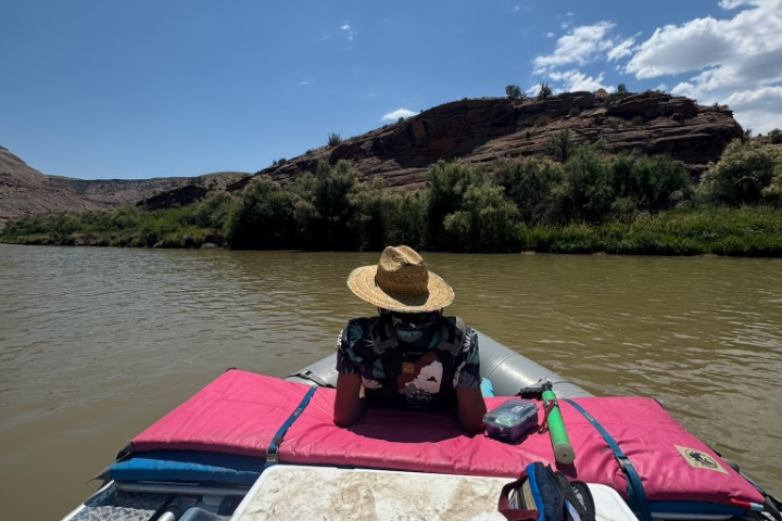 Person in a straw hat on a raft, facing river and hills under a clear blue sky.
